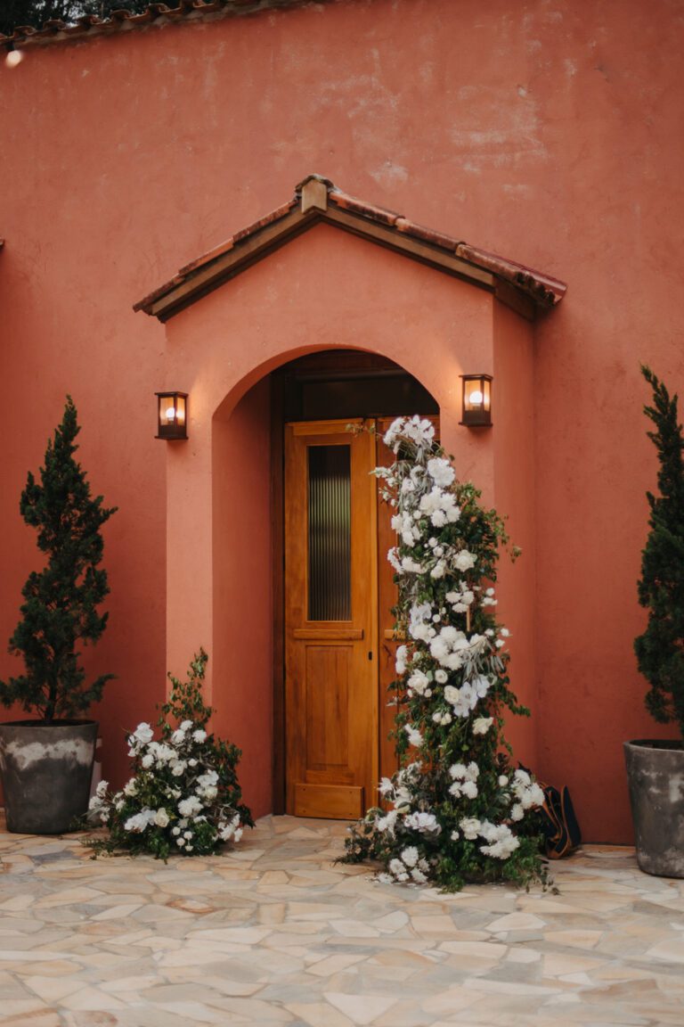 Uma porta de madeira em uma parede rosa, cercada por flores brancas e arbustos verdes, iluminada por luzes de parede.